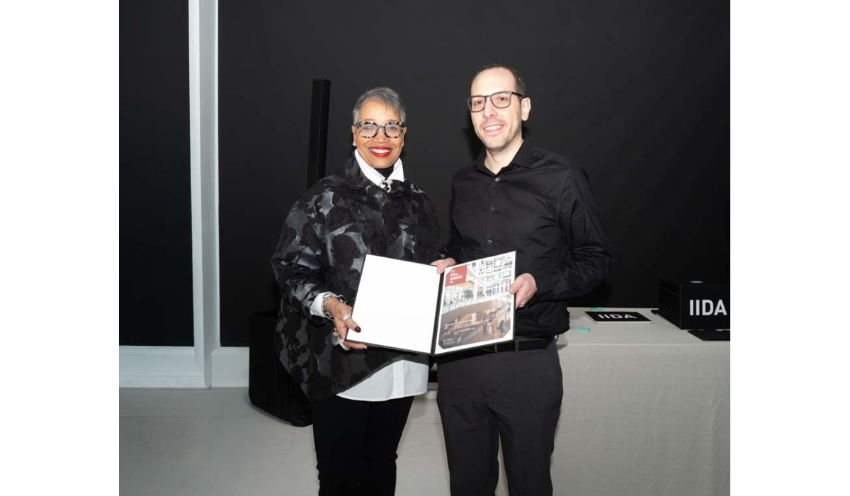Two individuals hold a magazine together, standing against a dark background. One person wears patterned glasses, while the other is dressed in black.