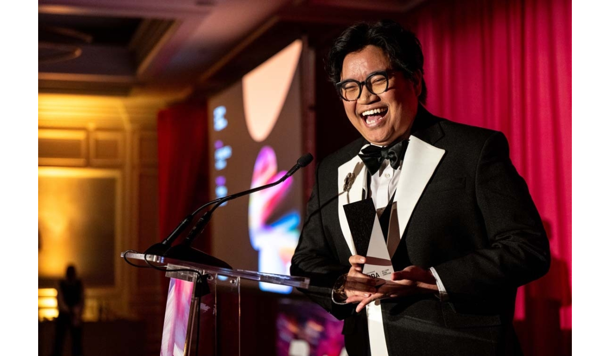 Smiling man in a black tuxedo and bow tie accepts a triangular award at a podium, red curtains and projection screen behind him.