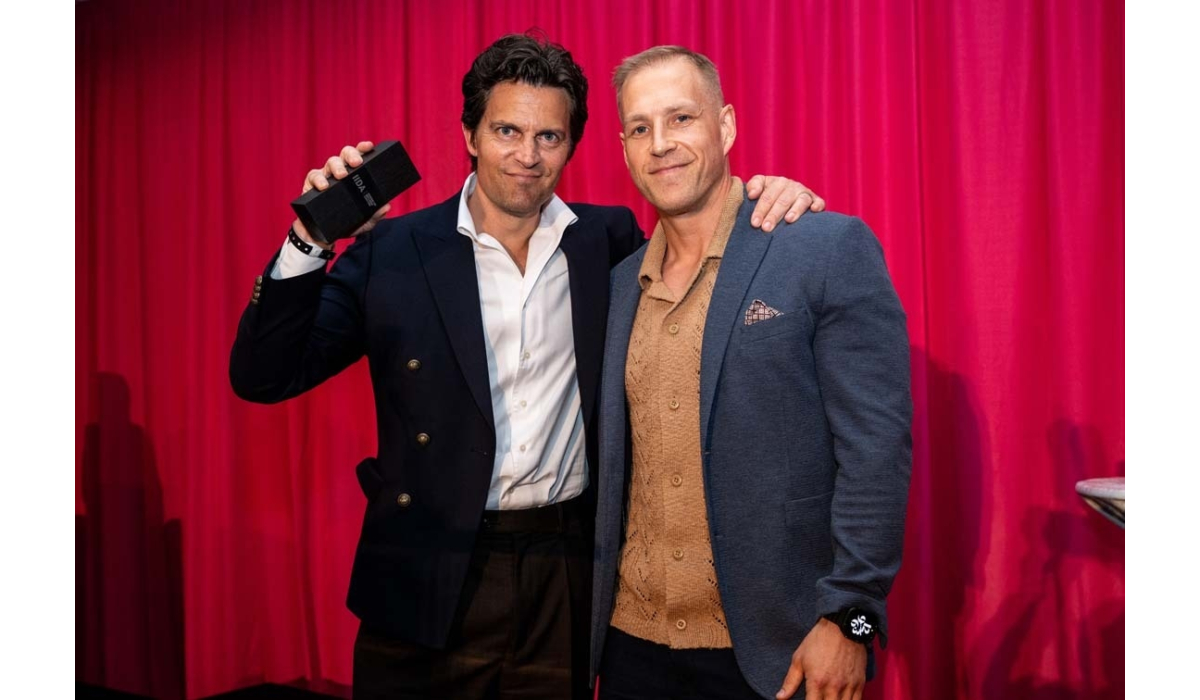 Two smiling men pose before a red curtain, one in a navy blazer holding a black award while the other rests a hand on his shoulder.