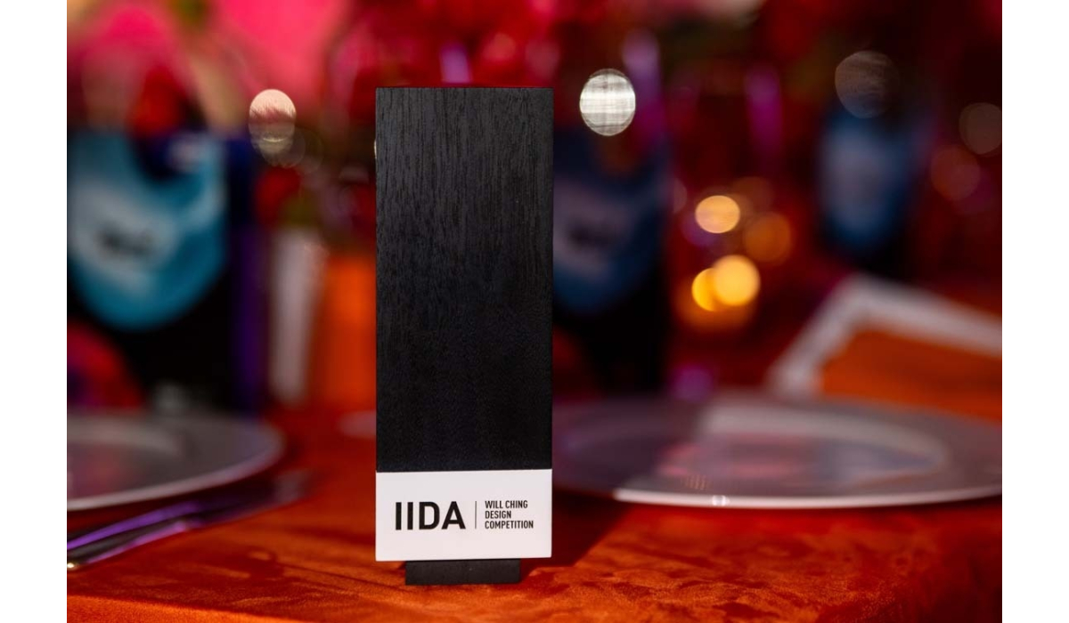Tall black award plaque labeled IIDA Will Ching Design Competition stands on an orange tablecloth amid colorful bokeh and tableware.