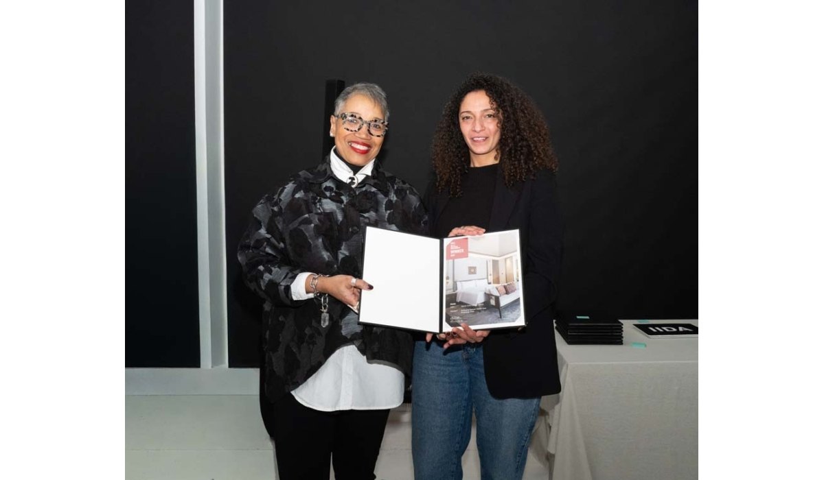 Two women stand together, smiling and holding an award book featuring a stylish interior design, with a modern backdrop.