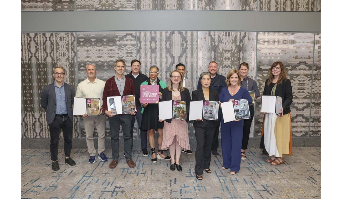 Twelve designers stand in a patterned conference room, smiling and holding open portfolios and an award plaque for library interior design.
