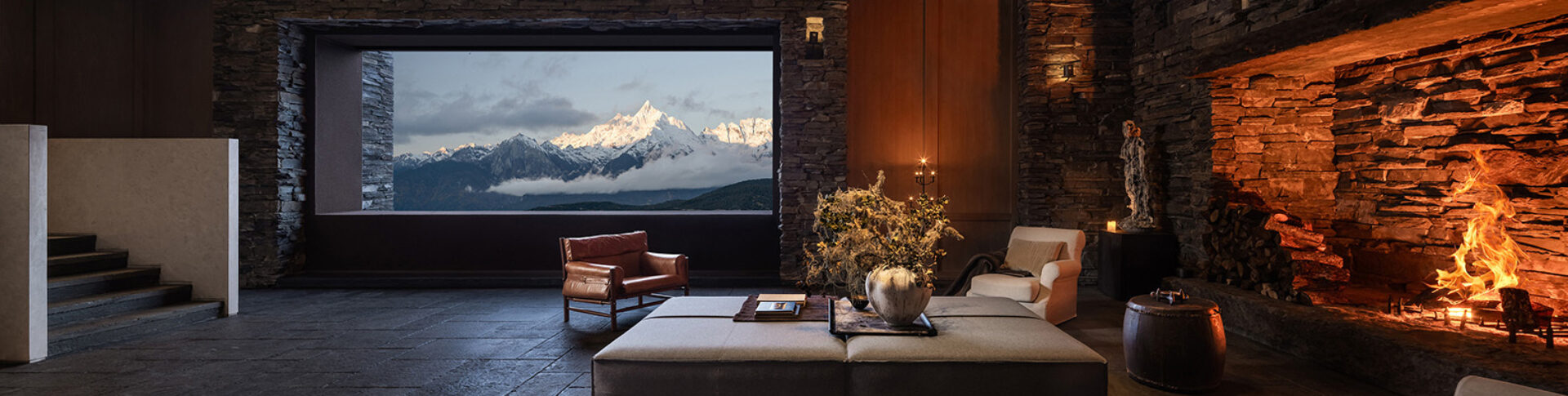Stone-walled mountain lounge with roaring fireplace, leather and upholstered chairs around an ottoman and a wide window framing snowy peaks.