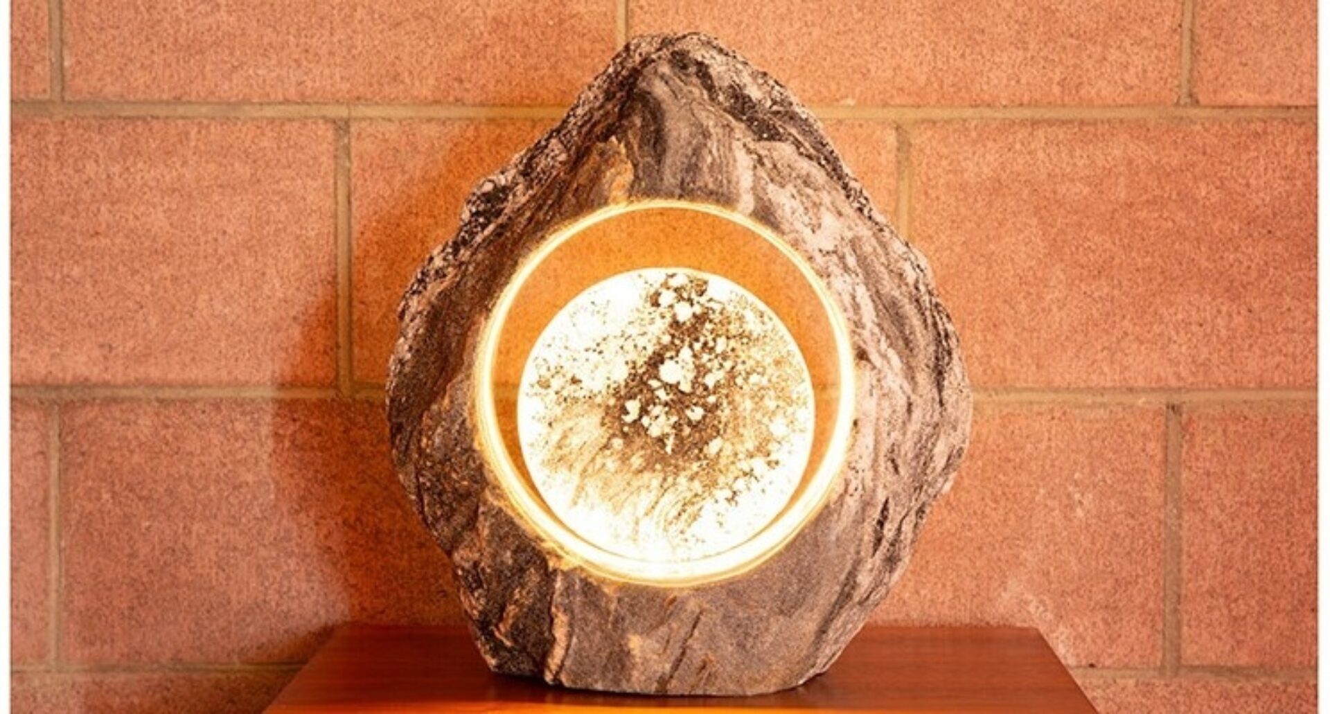 Rough volcanic stone lamp with a glowing circular alabaster center speckled with mineral veining, on a wooden table against a brick wall.