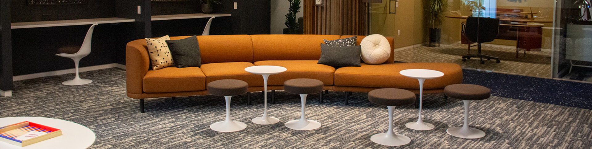 A modern lounge area features an orange sectional sofa with decorative pillows and several small, round white tables, enhancing comfort.