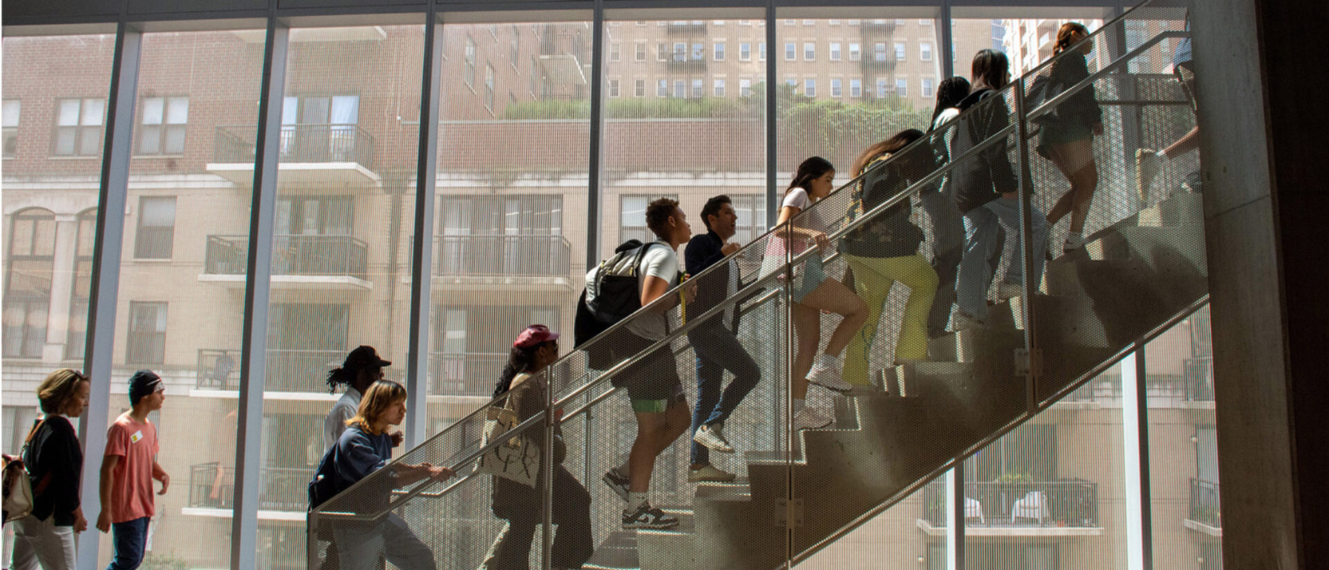 A diverse group of people ascends a modern staircase with large windows, creating a bright and inviting atmosphere.