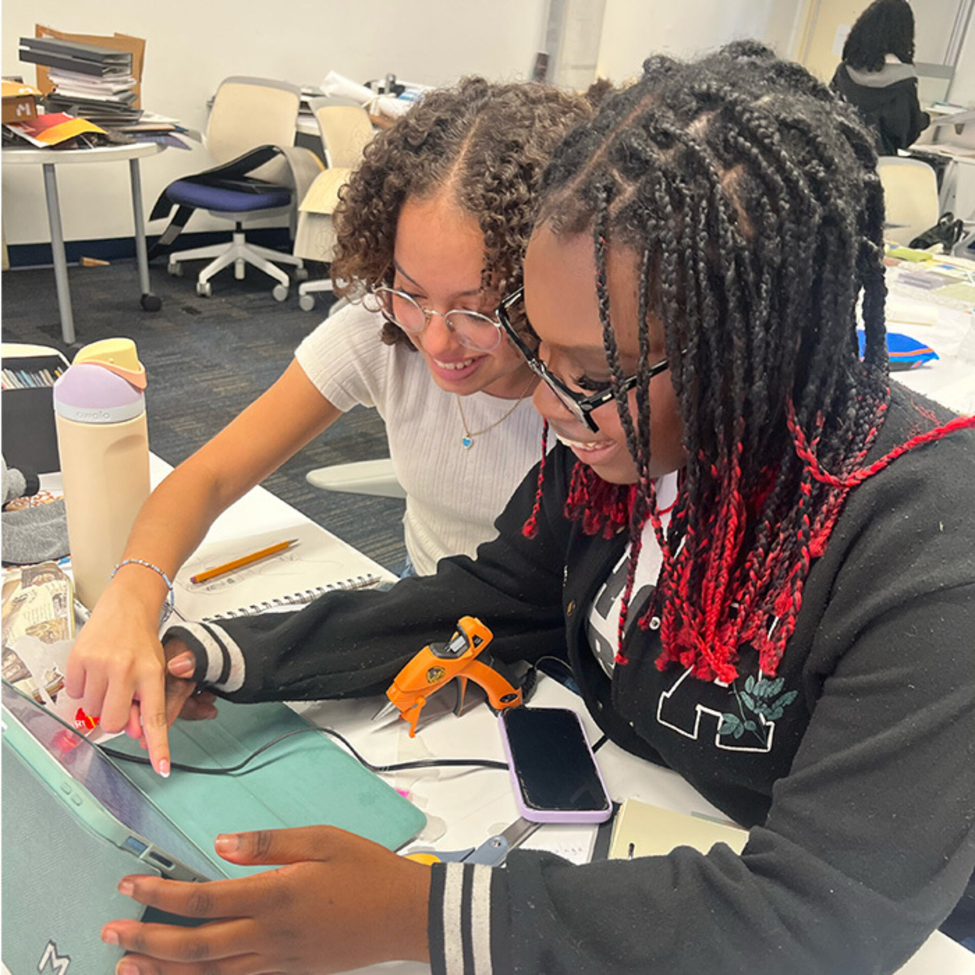 Two students collaborate at a table, focused on a tablet. One points to the screen, while the other examines it closely. Materials scattered around.