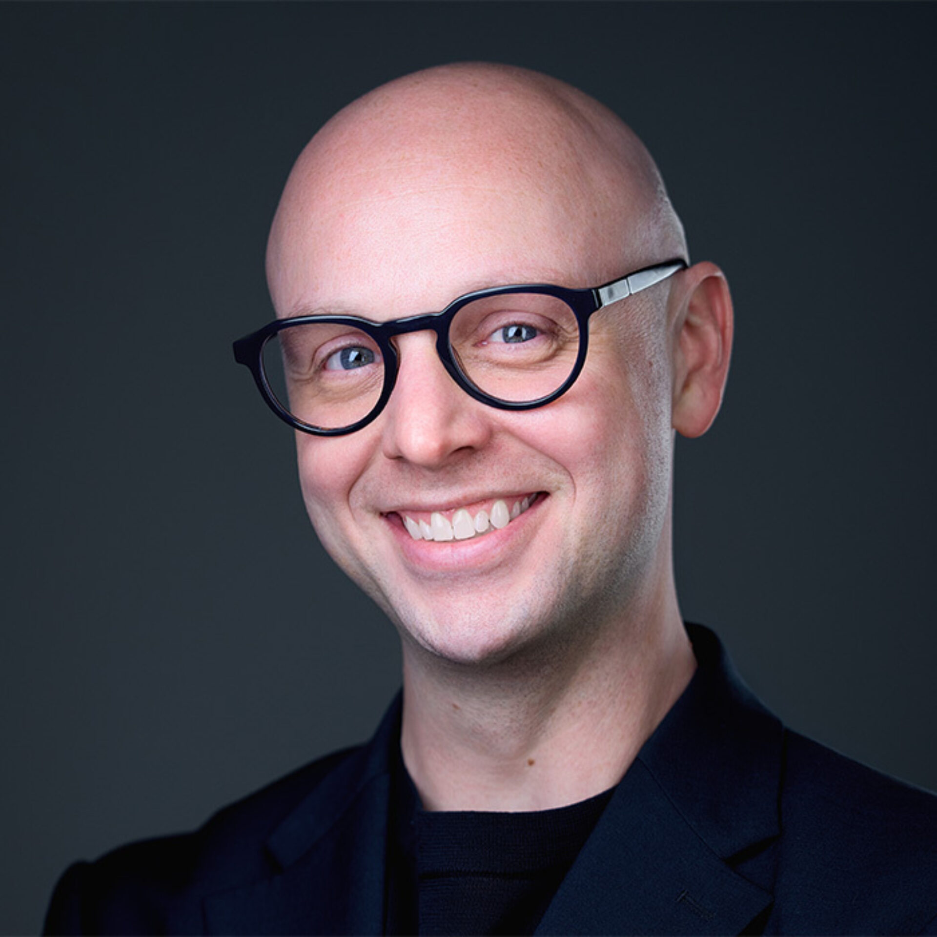 A smiling person with a bald head and glasses wears a black blazer, set against a dark background. The expression conveys warmth.