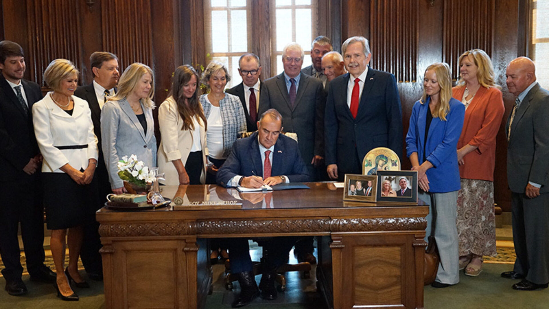 A man in a suit signs a document at an ornate wooden desk as a dozen dressed, smiling people stand behind him in a wood-paneled room.
