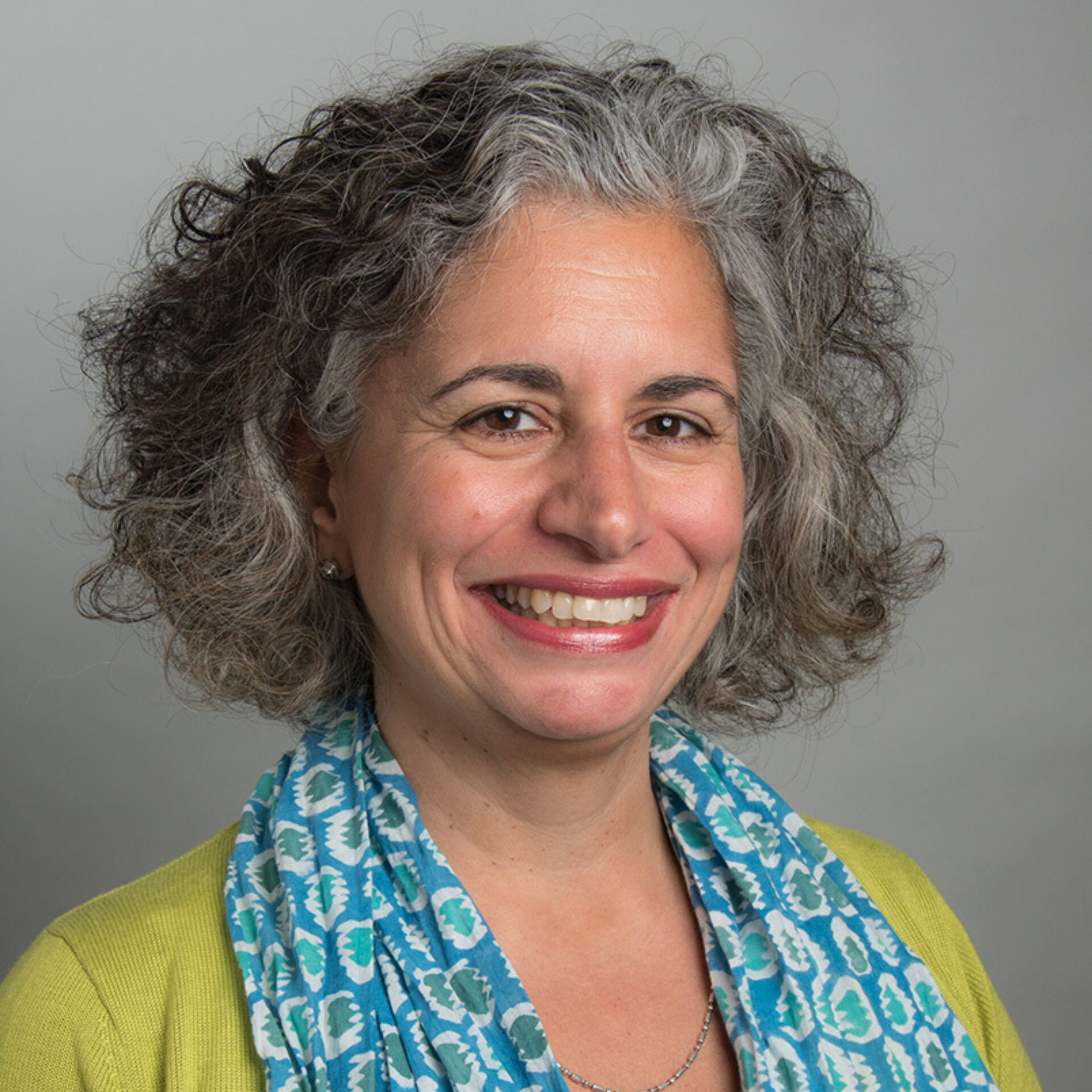 A smiling woman with curly gray hair wears a vibrant green blouse and a blue patterned scarf, posing in front of a neutral background.