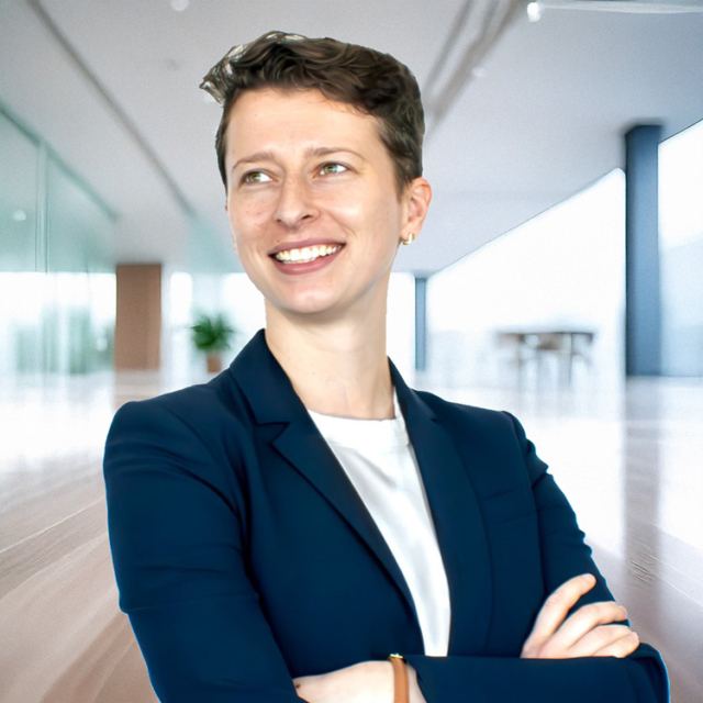A smiling woman with short hair, wearing a navy blazer and white shirt, stands confidently in a bright, modern office space.