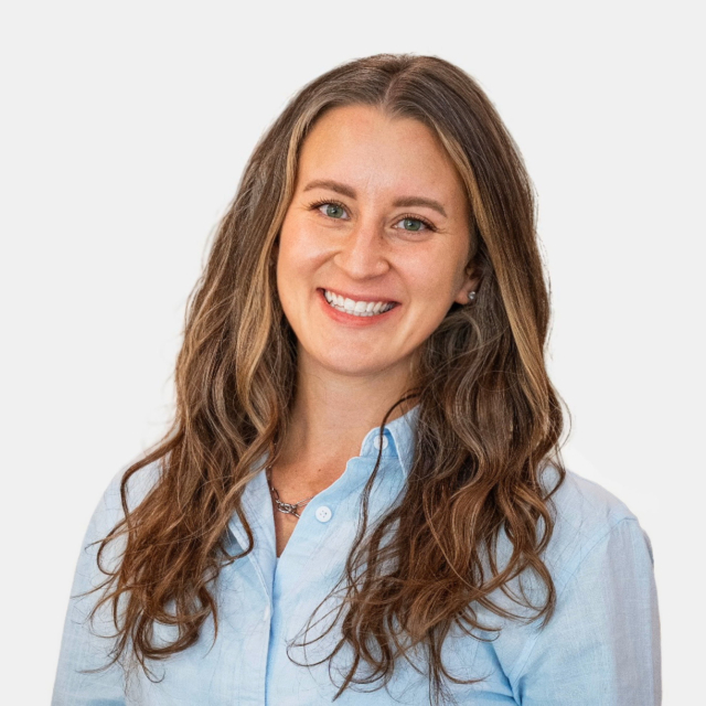 A woman with long, wavy brown hair and a bright smile wears a light blue shirt. She has a friendly expression against a white background.