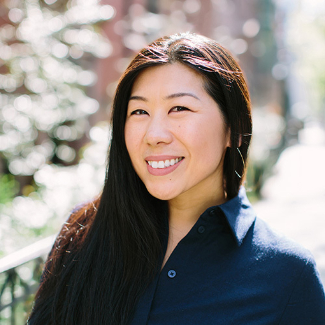 A woman with long dark hair smiles confidently while wearing a navy shirt, standing in a sunlit outdoor setting with blurred greenery.