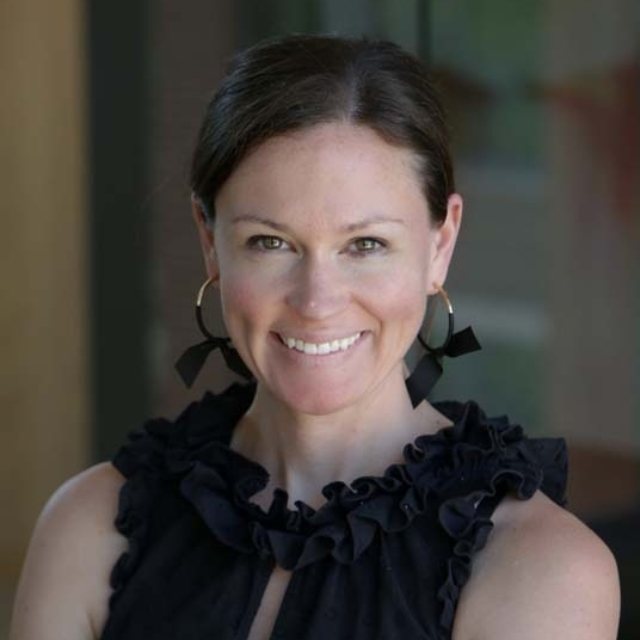 A smiling woman with brown hair, wearing a ruffled black top and hoop earrings, poses confidently against a blurred background.