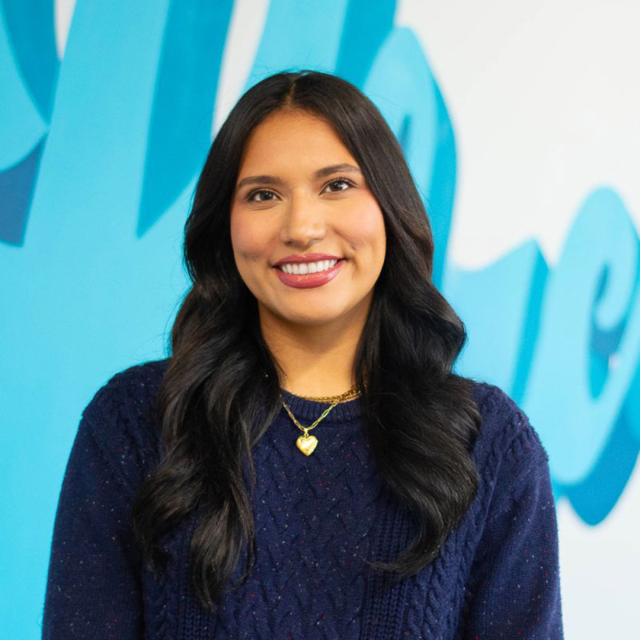 A young woman with long dark hair smiles brightly, wearing a navy sweater and a heart-shaped necklace against a blue backdrop.