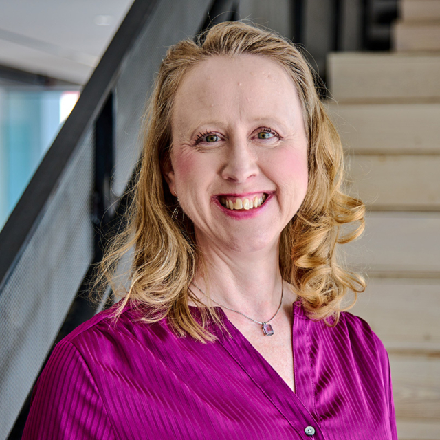 Smiling middle-aged woman with shoulder-length blonde curls wearing a magenta blouse and pendant necklace, posed by a metal stair rail.