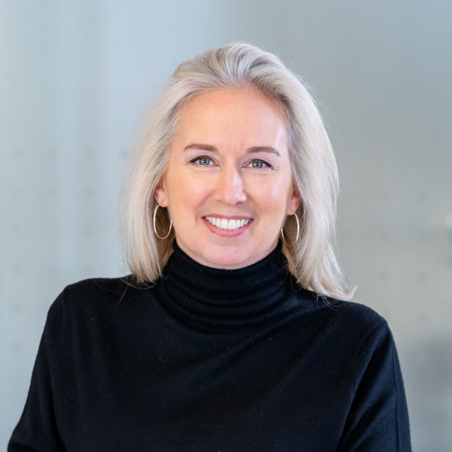 A woman with long, light blonde hair smiles warmly while wearing a black turtleneck. She stands against a softly blurred background.