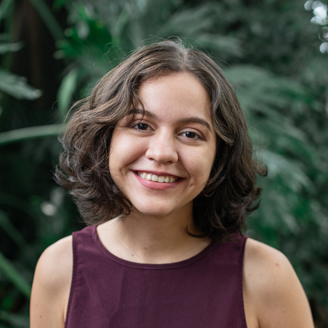 Smiling young woman with short wavy brown hair wearing a sleeveless burgundy top, posed against a soft-focus backdrop of green foliage.
