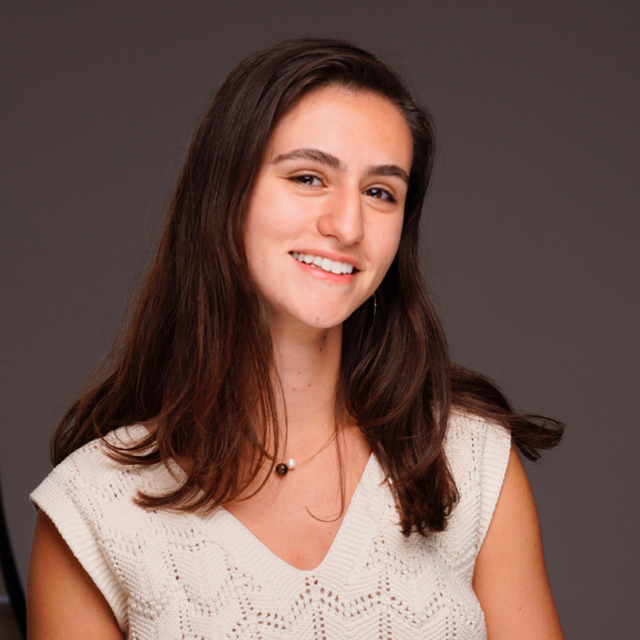 A young woman with long, dark hair smiles warmly, wearing a white knitted top against a neutral background. Her expression is friendly and approachable.