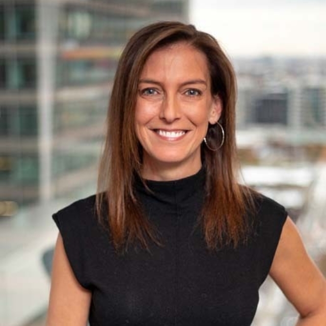 Smiling brown-haired woman in a sleeveless black top and hoop earring stands by an office window with blurred city buildings behind her.