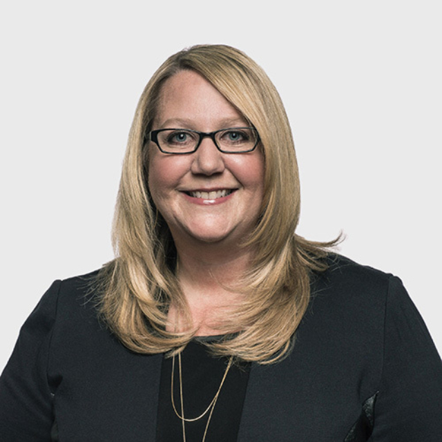 A smiling woman with long, blonde hair and glasses wears a black blazer, conveying a professional and approachable demeanor.