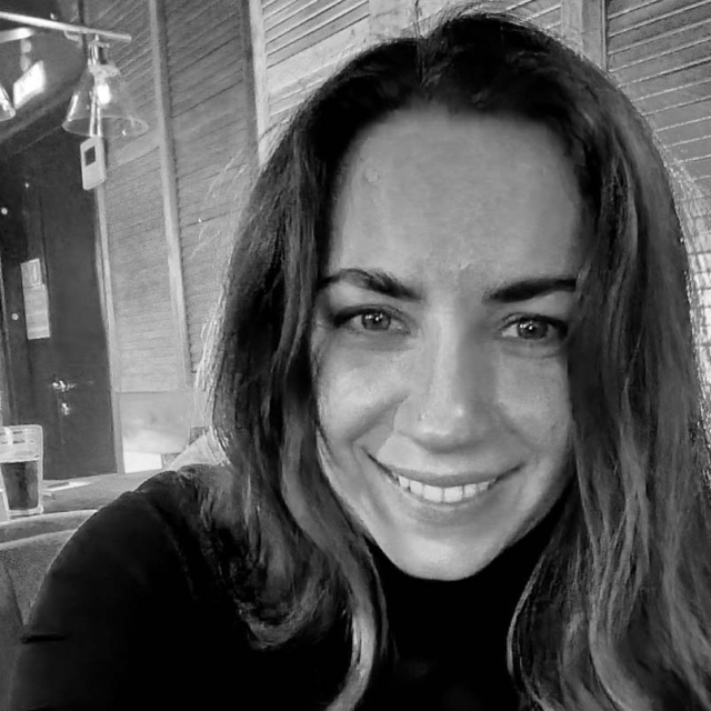 A woman with long, wavy hair smiles warmly at the camera, seated in a cozy, dimly-lit environment with a drink nearby.