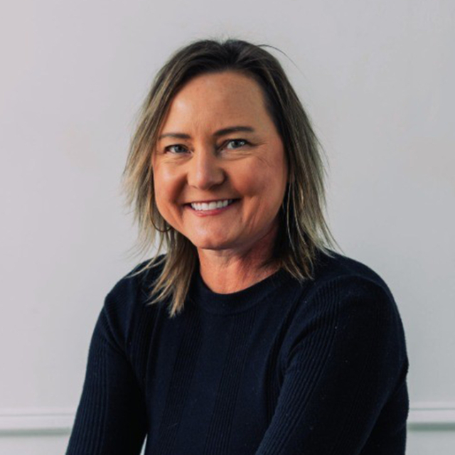 A woman with shoulder-length hair smiles, wearing a dark sweater against a light background, conveying a warm and approachable demeanor.