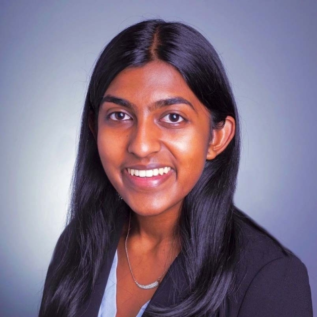 A young woman with long black hair and a bright smile wears a dark blazer over a light top, against a soft gray background.
