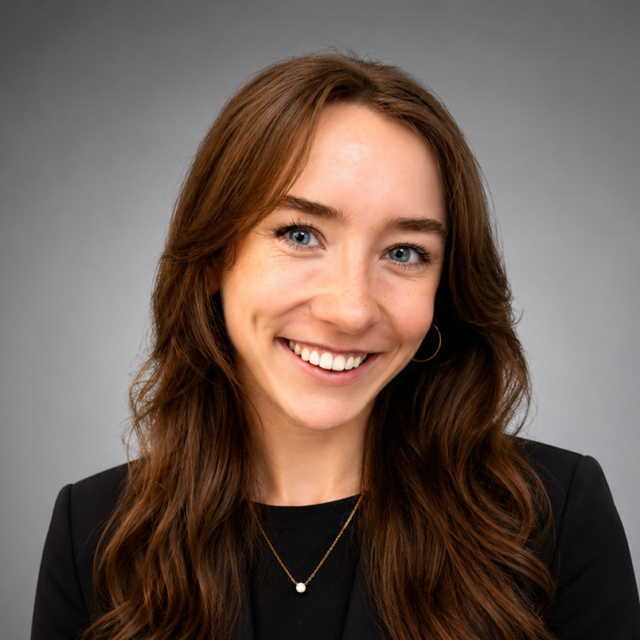 Smiling young woman with long wavy brown hair and blue eyes wearing a black blazer, small gold hoop earring and a delicate gold necklace.