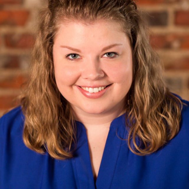 A smiling woman with curly hair, wearing a blue blouse, poses against a backdrop of rustic brick. Her bright eyes convey warmth.