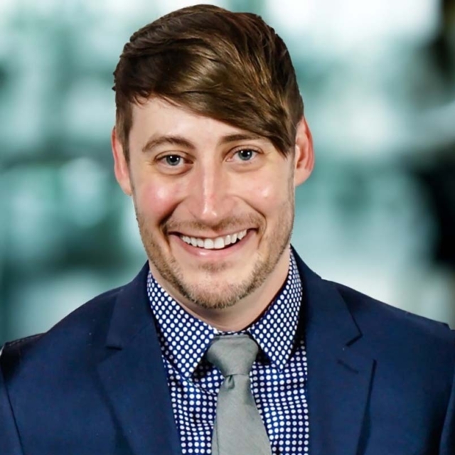 A smiling man with short brown hair and a well-groomed beard wears a dark suit, a polka dot shirt, and a tie in a blurred background.
