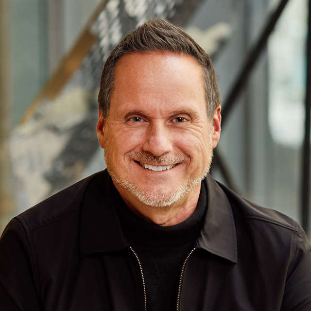 Smiling middle-aged man with short salt-and-pepper hair and trimmed beard, wearing black jacket and turtleneck, seated before glass stairs.