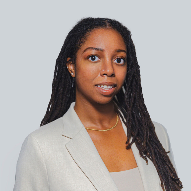 A woman with long, dark dreadlocks wears a light beige blazer and a simple gold necklace, smiling against a neutral background.