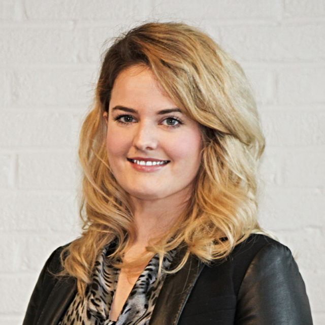 Smiling woman with wavy mid-length blonde hair, wearing a black leather jacket over gray patterned blouse, posed against a white brick wall.