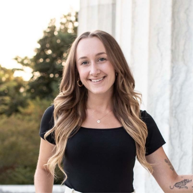 A smiling woman with long, wavy hair stands near a white marble wall, wearing a black top and light-colored pants. Natural light enhances the scene.