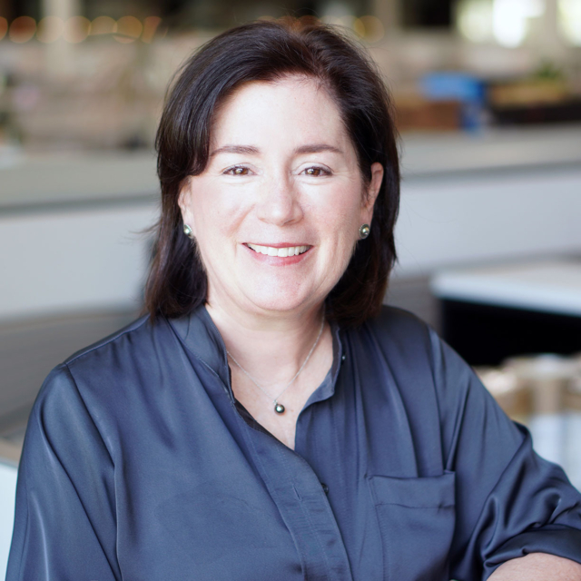 A smiling woman with dark hair, wearing a navy blouse and silver jewelry, poses in a well-lit, modern environment.