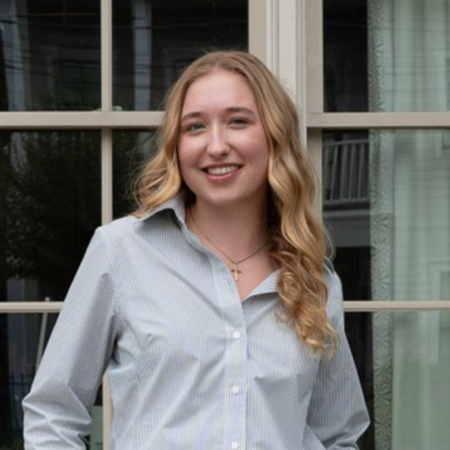 A young woman with wavy blond hair smiles confidently, wearing a light blue striped shirt, standing by a window adorned with greenery.