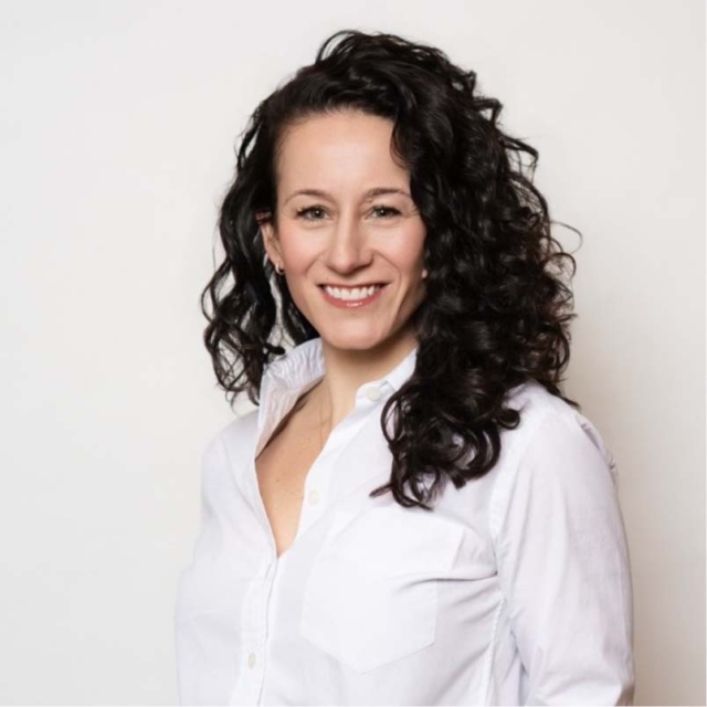 A woman with curly dark hair smiles confidently while wearing a white button-up shirt against a light background.