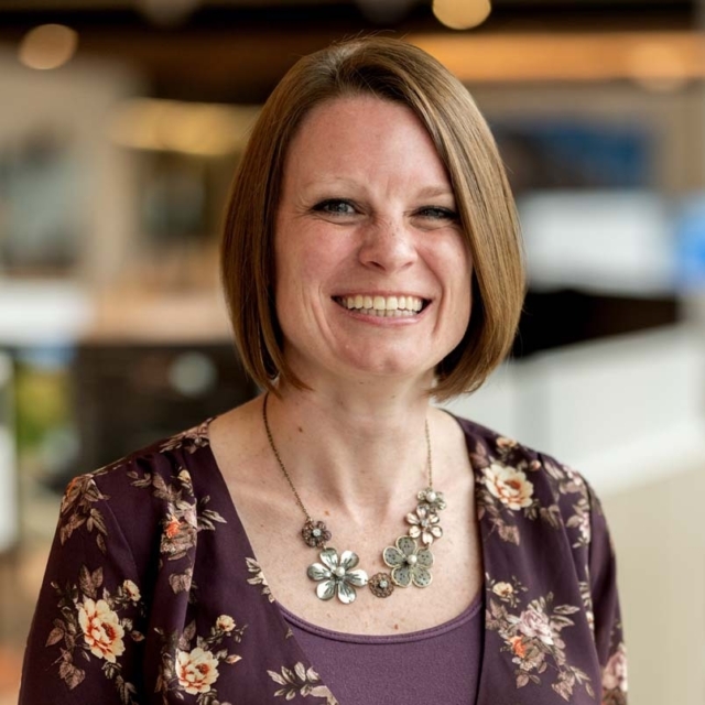 A woman with shoulder-length brown hair smiles warmly, wearing a floral blouse and a statement necklace, in a modern indoor setting.