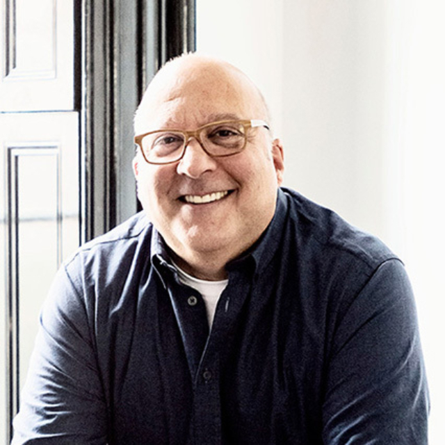 Bald, bespectacled smiling man in navy button-up and white tee sits by a tall black-framed window, warm natural light on his face.