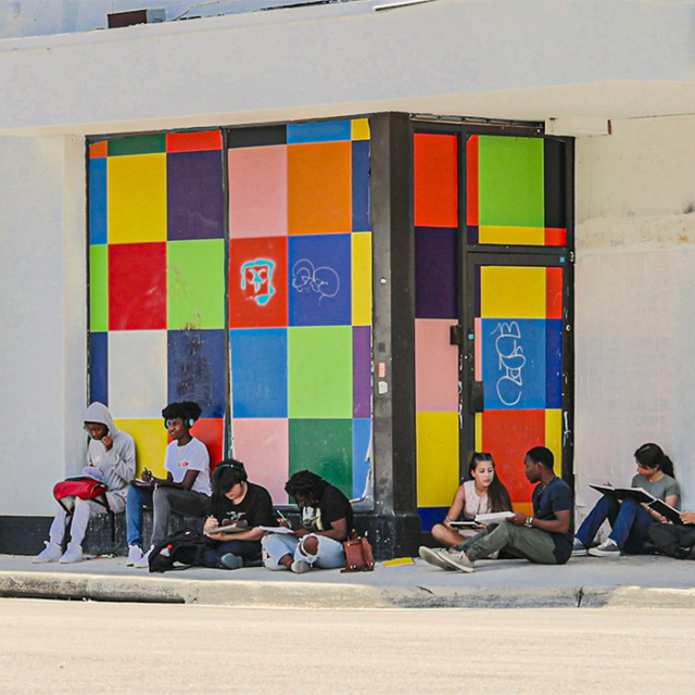 A vibrant mural with colorful squares serves as a backdrop for a diverse group of people seated on the sidewalk, some sketching.