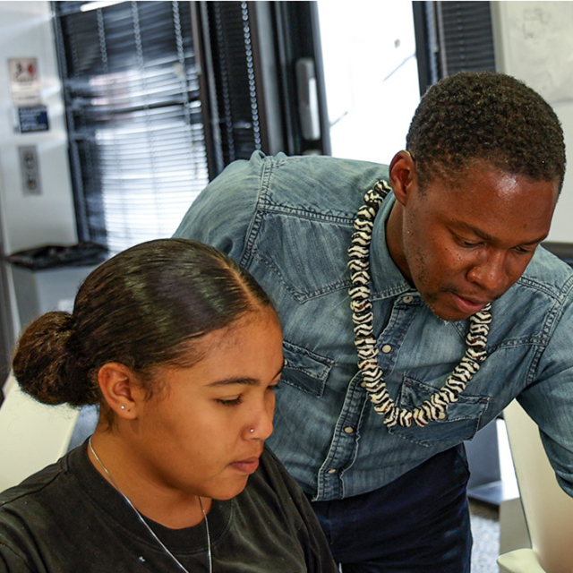 A young woman focuses on her work while a man leans over to provide guidance, both engaged in a creative learning environment.