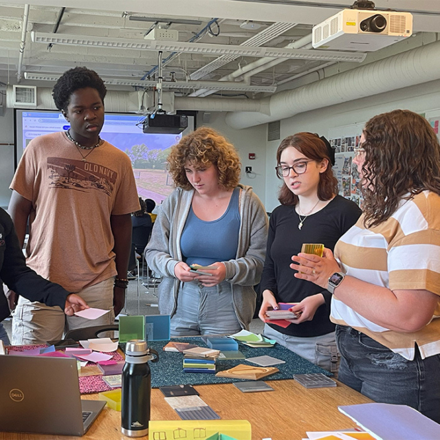 A diverse group of students collaborate at a table filled with color swatches and materials, engaged in a design project.