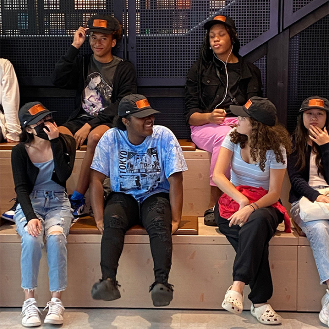 A diverse group of young people sits on a wooden bench, wearing matching black caps, enjoying each other's company and laughter.