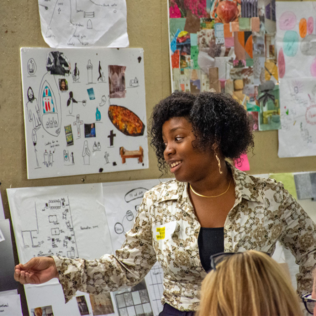 A person with curly hair, wearing a patterned shirt and necklace, gestures while speaking in front of a wall filled with various art boards.