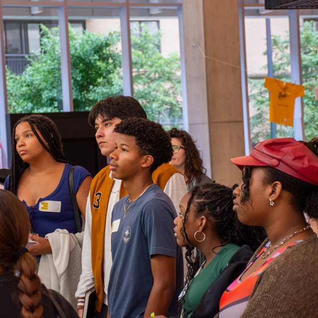 A diverse group of young people attentively listens to a presentation in a well-lit space, surrounded by greenery and casual decor.