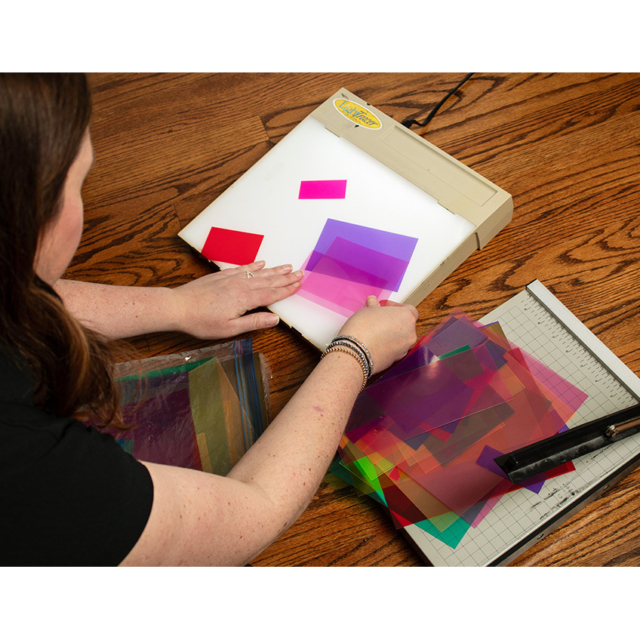 Person arranging translucent colored gel squares on a lit lightbox, with a stack of vibrant gels and a paper cutter on a wooden table.