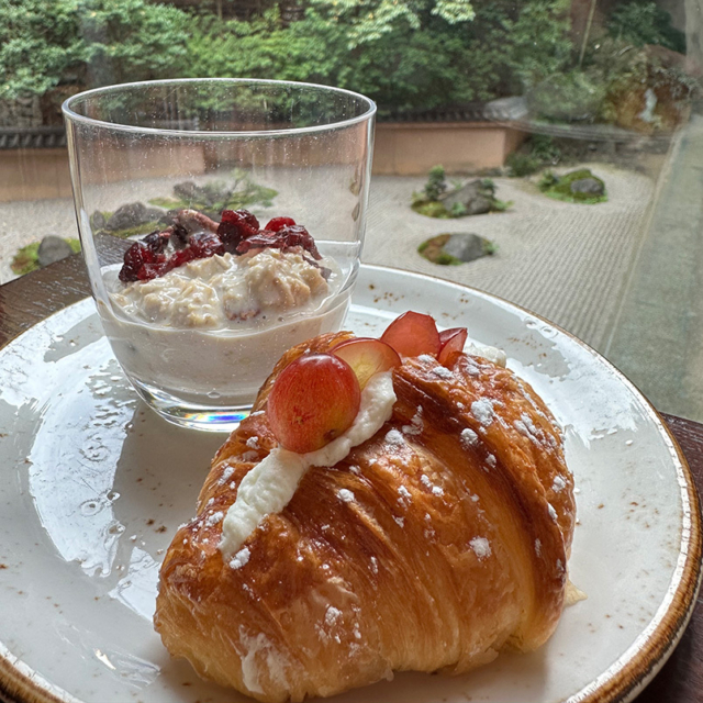 A buttery croissant garnished with red fruit and cream sits beside a glass of yogurt topped with berries, against a serene garden backdrop. (Image courtesy of Lori Mukoyama)