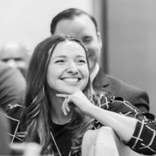 A smiling woman rests her chin on her hand, engaged in conversation amid a group of people, conveying a sense of warmth and joy.