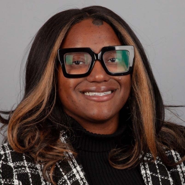 A person with long, dark hair and glasses smiles, wearing a black turtleneck and a patterned jacket, against a neutral background.